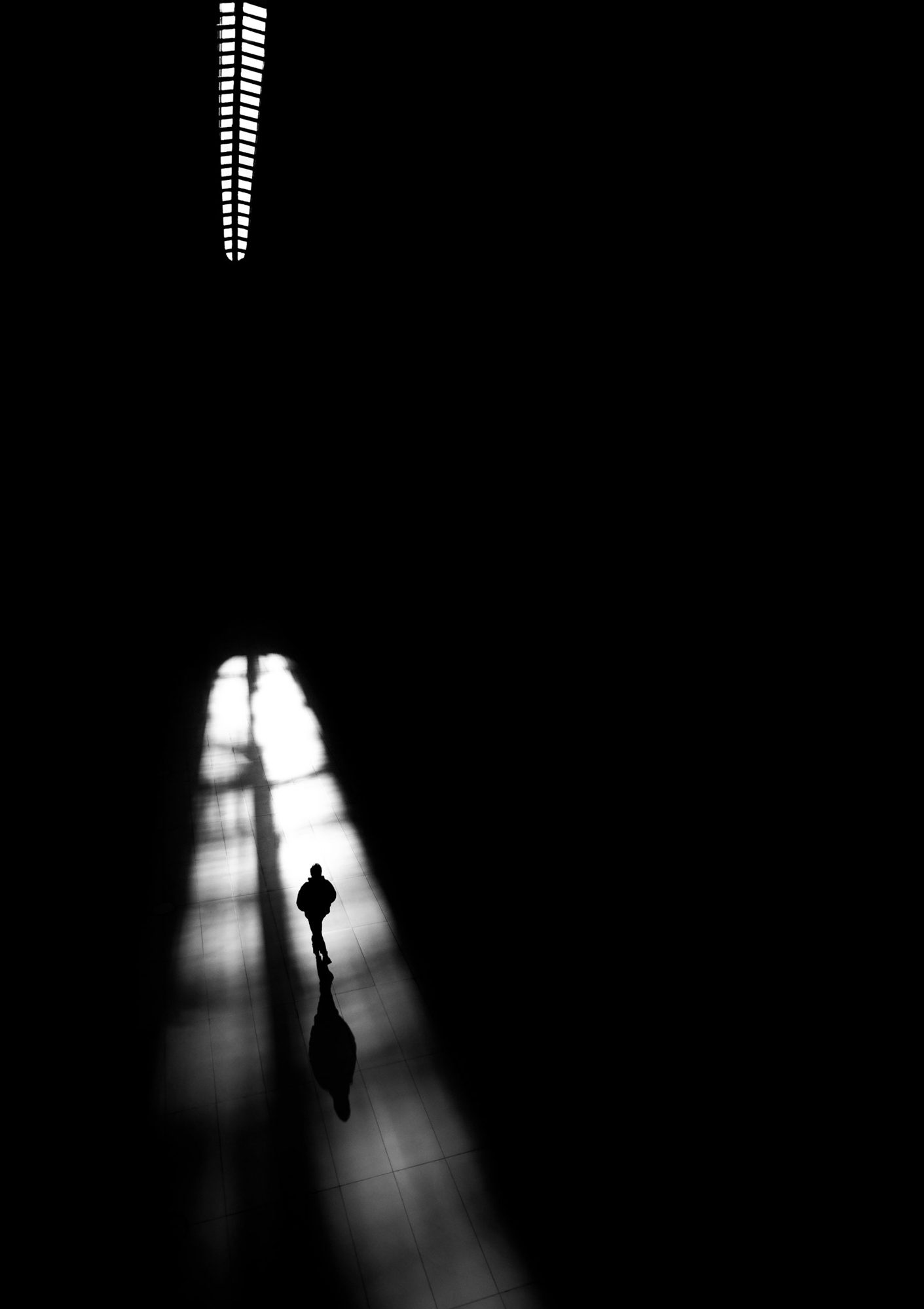 Black-and-white photograph: A lone figure walks in a shaft of light across a dark hall, seen from above