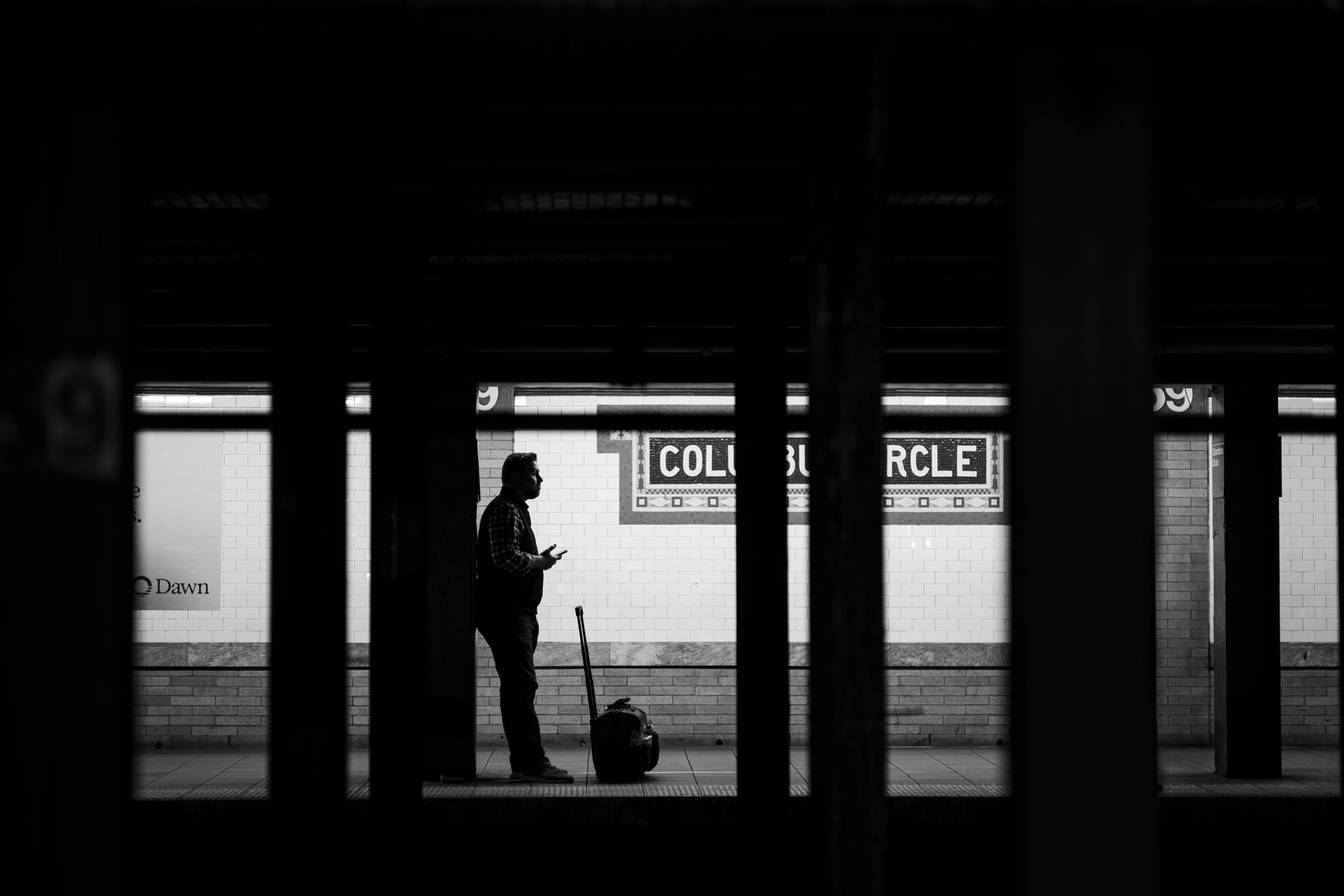 Black-and-white photograph: A man with a rolling bag waits on a subway platform, framed between dark pillars at Columbus Circle